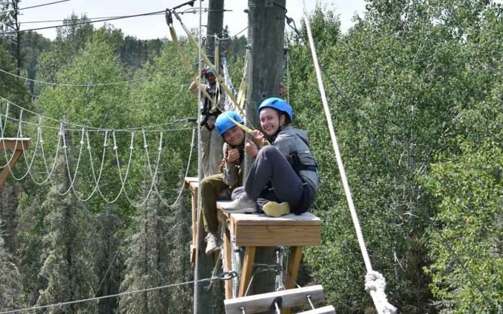 two students smile while sitting atop a platform on a ropes course 